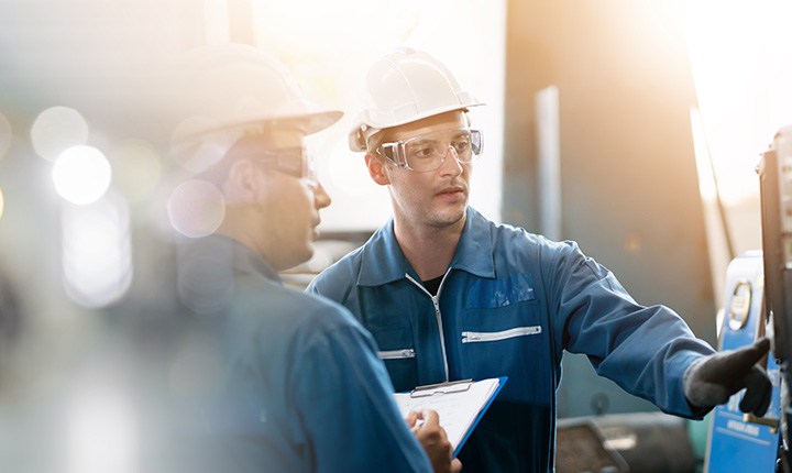 Due ingegneri in tenuta protettiva discutono di attrezzature mentre uno indica una macchina e l'altro tiene una cartellina.