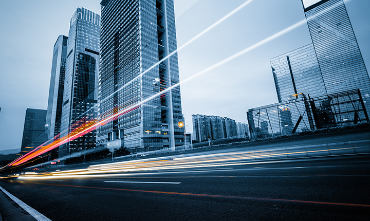 Alti grattacieli moderni con scie luminose sfocate di veicoli in movimento su un'ampia strada cittadina al tramonto.