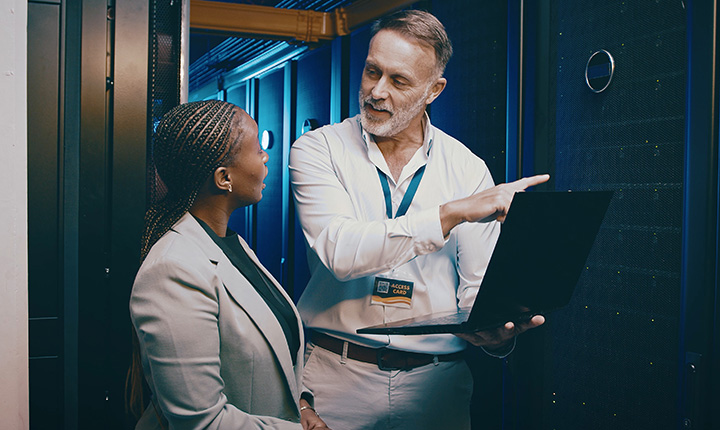 Due professionisti si trovano in una sala server; uno tiene in mano un computer portatile e gesticola mentre spiega qualcosa all'altro.