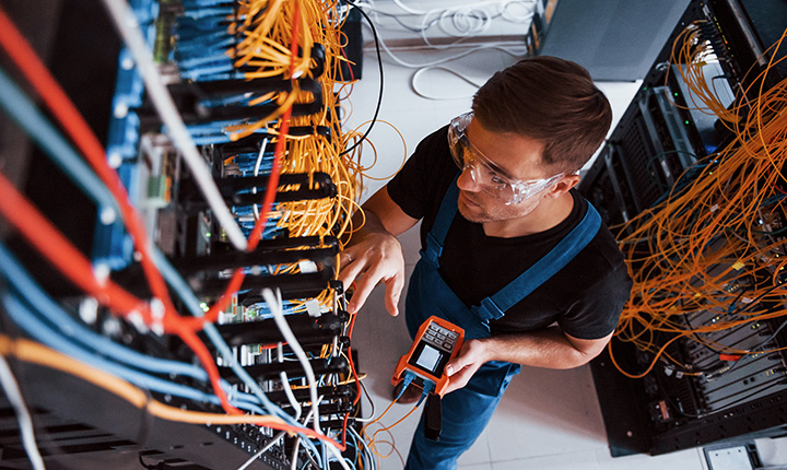Un tecnico con occhiali di sicurezza testa i cavi con un dispositivo portatile in una sala server piena di cavi di rete.