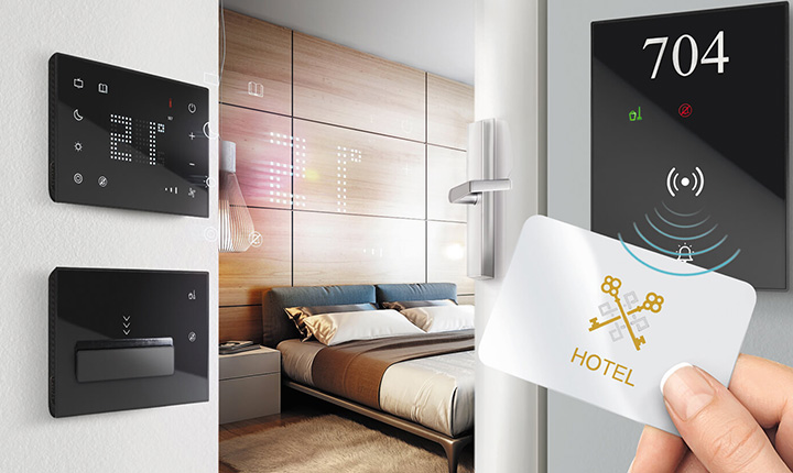 Una mano tiene una chiave d'albergo vicino a un pannello digitale della camera; sullo sfondo è visibile una moderna camera d'albergo.