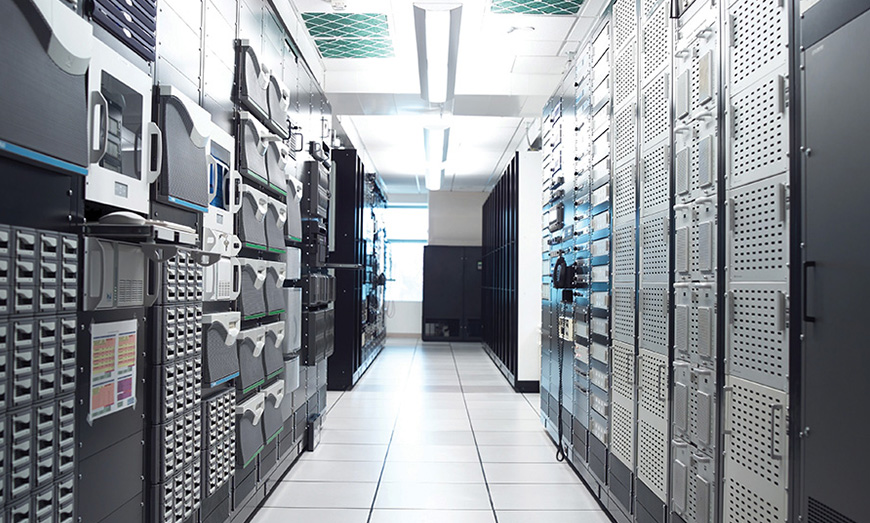 Un centro dati ben illuminato con file di rack di server e apparecchiature informatiche allineate su entrambi i lati di un corridoio.