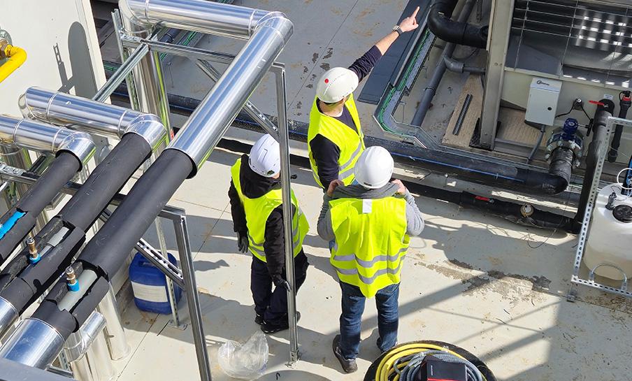 Tre operai con giubbotto ed elmetto di sicurezza ispezionano tubi e attrezzature industriali in un cantiere all'aperto.