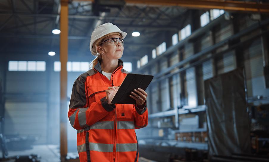 Una persona in tenuta di sicurezza e cappello rigido si trova in un magazzino industriale e scrive su un tablet.