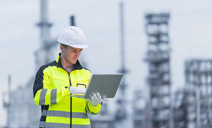 Un operaio edile con giacca ad alta visibilità e elmetto utilizza un computer portatile in un sito industriale.