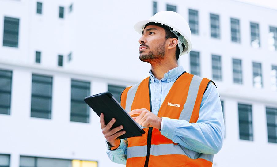 Un uomo con elmetto e giubbotto di sicurezza arancione utilizza un tablet mentre si trova all'esterno di un edificio.